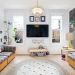 Bright living room with a wall-mounted TV between two windows, flanked by colorful striped couches and a round rug at center.