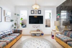 Bright living room with a wall-mounted TV between two windows, flanked by colorful striped couches and a round rug at center.